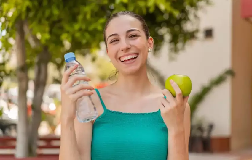 mulher branca sorrindo segurando uma maça e uma garrafa de água para contribuir na hidratação para seu emagrecimento