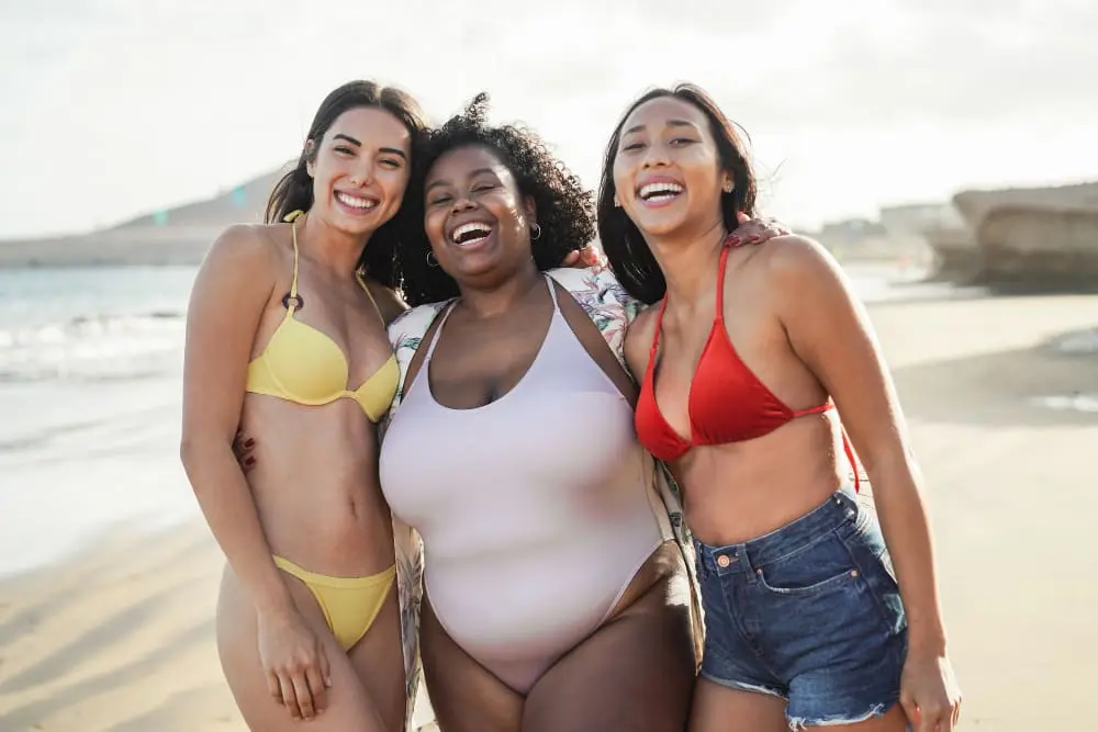 tres-mulheres-abracadas-na-praia-com-biquinis-vermelho-branco-e-amarelo-sorrindo-com-seus-corpos-de-verao